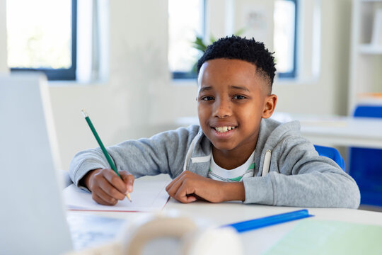 African American schoolboy sitting in classroom at desk writing in notebook with green pencil