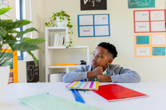 African American child sitting at white desk in classroom, resting chin, gazing right at notebooks