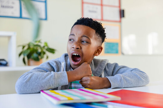 African American preteen in gray hoodie yawning, leaning on desk in classroom with bright notebooks