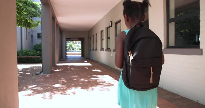 Child is walking in shaded walkway drawn by sunlit courtyard, glancing right and adjusting strap