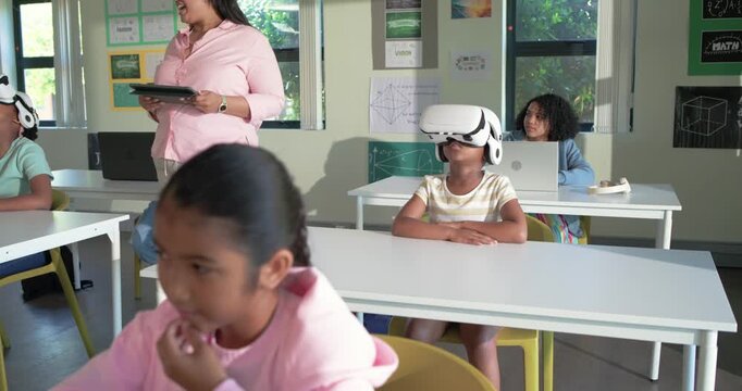 Teacher checking tablet, walking aisle, monitoring students in white VR headsets raising hand