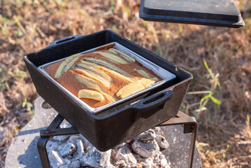Freshly baked banana cake with banana slice toppings in a mold heating over hot charcoal embers. Concept of outdoor cooking, camping lifestyle, and homemade dessert.