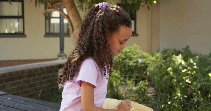 Female child taking pad from yellow backpack and drawing panels on bench while peer eating snack