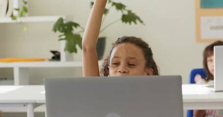 African American girl child peeking over laptop, raising hand in classroom for teacher attention