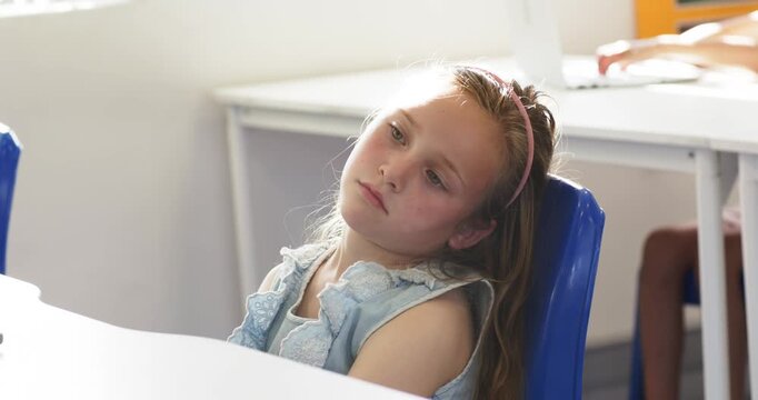 Girl child sitting at white desk in classroom, tilting head left while contemplating window light