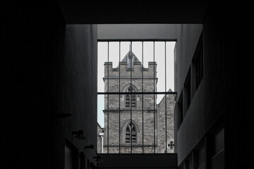The stone tower of St Ebbe's Church in Oxford, framed by the dark interior and grid-like bars of a modern adjacent building of the Westgate Centre.