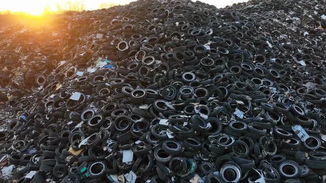 A large pile of discarded tires is visibly stacked under a setting sun, showcasing the overwhelming amount of waste. The discarded tires accumulate, creating an unsettling sight in the environment.