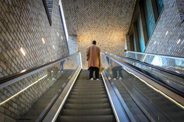 A view of a person in a tan overcoat ascending a modern escalator within the Westgate Shopping Centre in Oxford. The escalator leads to the roof with a beautiful panoramic view of the city.