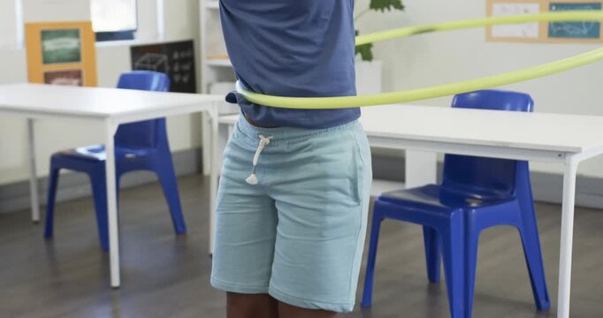 Child is entering frame and spinning yellow hula hoop at waist in classroom, practicing balance