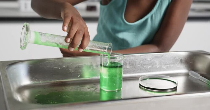 Child is tilting cylinder, pouring green liquid into beaker on tray making foam overflow for demo