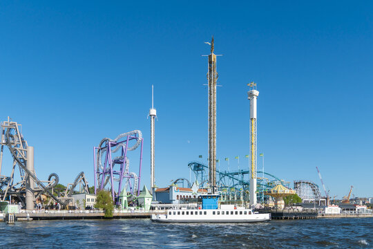 View of amusement park rides punctuate the horizon, contrasting against the serene blue sky and water, with a white boat sailing by, Stockholm, Stockholm County, Sweden.