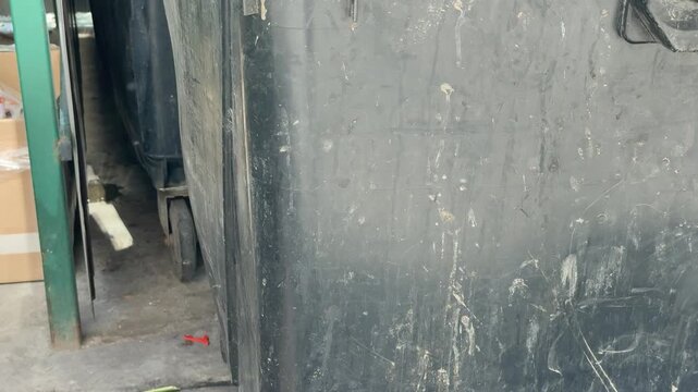 Weathered black dumpster under corrugated shed. Closeup view of large wheeled plastic container with chipped paint and rusted casters, splattered with white residue and grime,