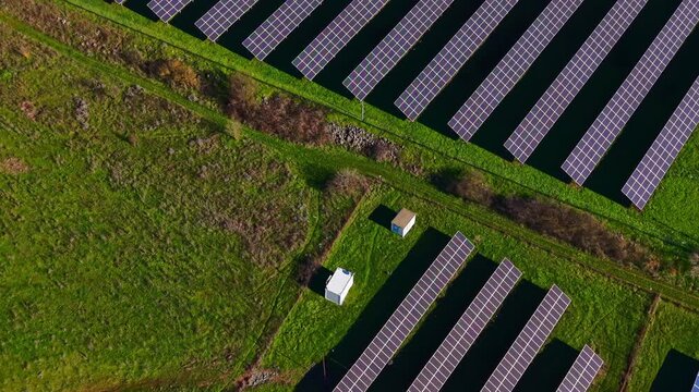 A large area of land shows numerous solar panels arranged in rows. A small building is located in the middle of the solar field. This scene shows renewable energy production in action.