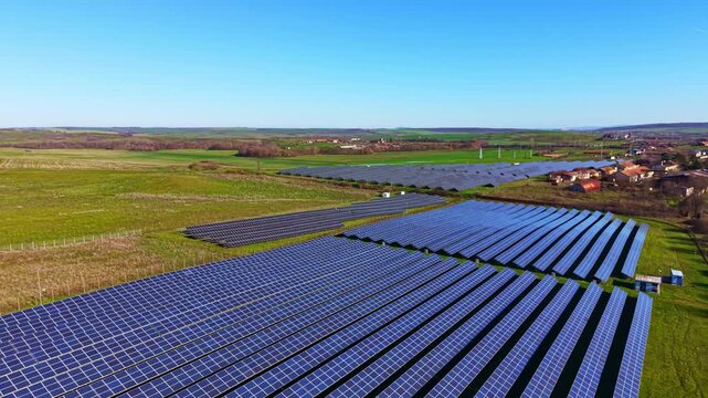 Solar panels are arranged in rows across a green field. A clear blue sky is overhead while some buildings are visible in the background. The sun is shining bright.
