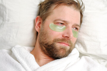Handsome 40s man with cosmetic patches under the eyes. Male takes care of his face skin in the morning. He applying green collagen patches. Facial treatment. Cosmetology beauty spa. Eye mask, wellfare