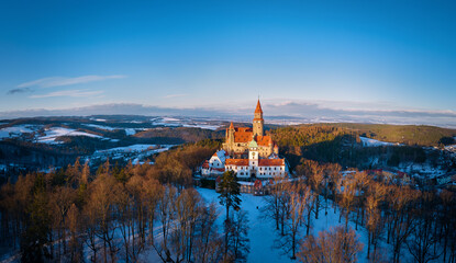 Panoramic aerial view of Bouzov Castle rising above snowy forest and rolling Czech countryside at golden hour