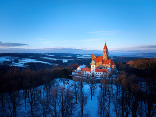 Panoramic aerial view of Bouzov Castle rising above snowy forest and rolling Czech countryside at golden hour