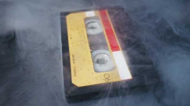 Old audio cassette tape with a yellowed label lies on a dark surface as thick white smoke swirls around it. A slow camera pan reveals the retro object, creating a nostalgic and mysterious atmosphere.