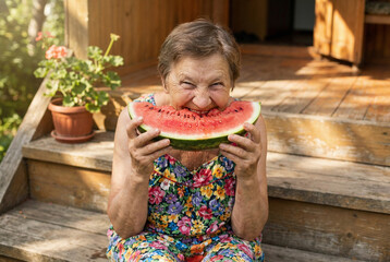 A cheerful senior woman playfully bites a juicy watermelon slice, sitting on a wooden porch. A funny, happy moment of summer vacation and simple pleasure.