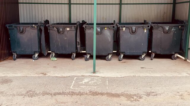 black dumpsters on concrete alley, closeup of worn caster wheels, scuffed lids and chipped paint, scattered debris and small puddles on pavement, industrial fence background,