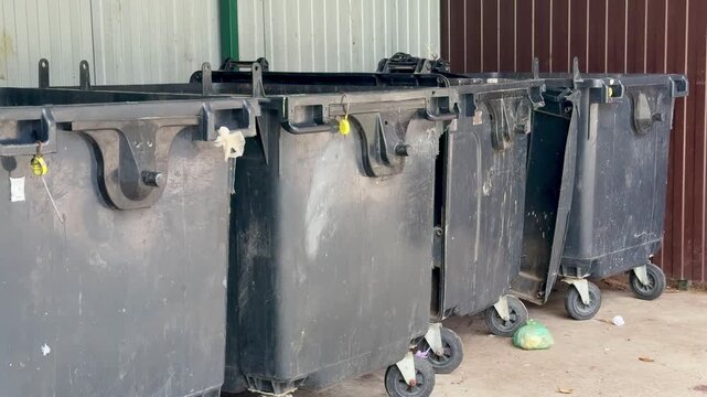 black dumpsters on concrete alley, closeup of worn caster wheels, scuffed lids and chipped paint, scattered debris and small puddles on pavement, industrial fence background,