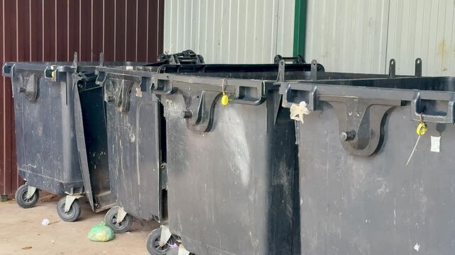 black dumpsters on concrete alley, closeup of worn caster wheels, scuffed lids and chipped paint, scattered debris and small puddles on pavement, industrial fence background,