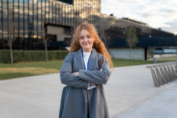 Fototapeta premium Red haired businesswoman standing with crossed arms in front of corporate building, showing confidence and professionalism