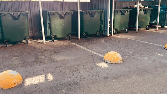 Dirty black green dumpsters on concrete inside metal shed under green supports exterior splattered with paint and mud, caster wheels stained, puddles and debris on rough floor closeup