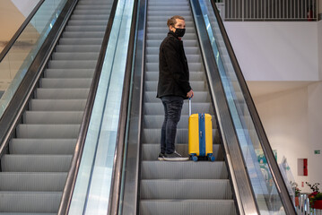 Man with face mask at the airport with a rolling suitcase on escalator