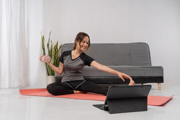 Asian trainer holds small dumbbell on mat, seated twist for tone and mobility with laptop for virtual coaching, compact home