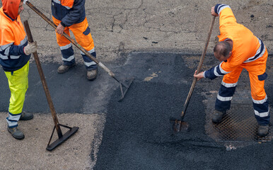 Workers in orange safety uniforms spreading hot asphalt with manual tools during road repair. High angle view of urban infrastructure maintenance and paving process. © peuceta