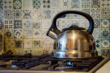 Stainless steel kettle heating on gas stove, against decorative tile wall, creating warm kitchen atmosphere.