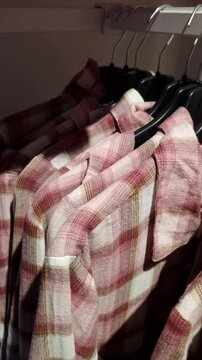 Pink and white plaid flannel shirts hang in a row on black hangers on a retail clothing rack. The camera slowly pans across the textured fabric in a softly lit environment.