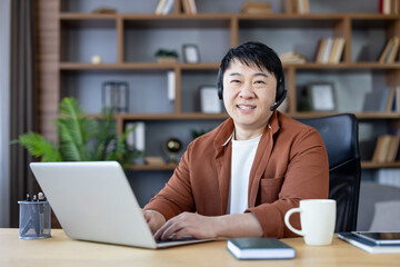Asian man smiling, wearing a headset with microphone, sitting at a desk and typing on a laptop, working or communicating online from a modern home office