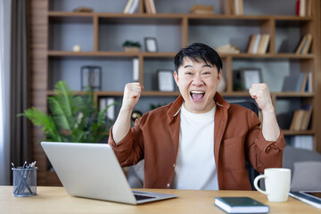 Asian man at home office desk cheering with clenched fists in front of laptop, celebrating work or study success, joyful, excited expression, casual modern interior