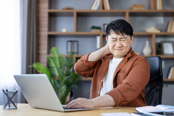 Asian man experiencing severe neck discomfort and pain, rubbing his sore neck while working from home on a laptop, highlighting occupational health issues and poor posture