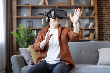 Asian man wearing a vr headset and headphones, sitting on a sofa in his living room, actively gesturing while interacting with an immersive metaverse experience or playing a video game
