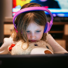 Young girl wearing headphones watching tablet while hugging teddy bear at home in evening