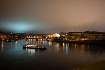 Fototapeta premium Prague city at night with illuminated buildings and a boat on the river Vltava during December. Cityscape of the historical capital of Bohemia in the Czech Republic.