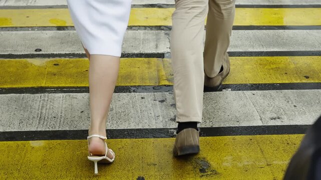 Close-up of a couple walking across a yellow-striped crosswalk, showing a woman in a white dress and high-heeled sandals beside a man in beige trousers and brown shoes