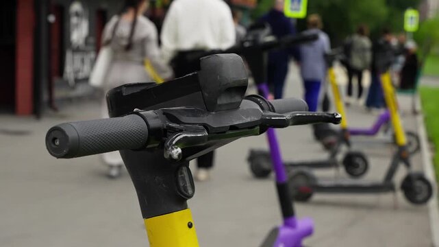 Detailed close-up of an electric scooter handlebar on a city sidewalk, with multiple shared scooters lined up and pedestrians moving in the blurred background during daytime.