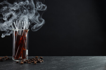 Glass test tube with coffee and beans on black table against dark background with smoke, space for text