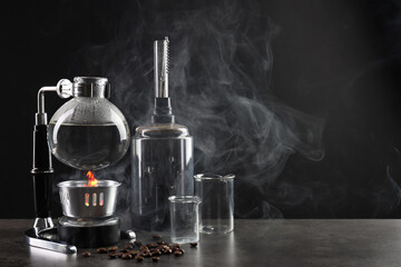 Vacuum coffeemaker, other equipment for making coffee and beans on grey table against dark background with smoke, space for text