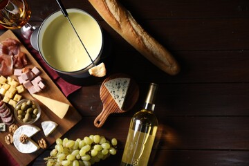 Fondue pot (caquelon) with melted cheese, wine and snacks on wooden table, flat lay. Space for text