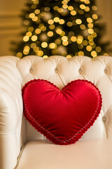 A red heart-shaped pillow on a white luxury sofa against a blurred backdrop of a glowing Christmas tree. Vertical template for Valentine's Day, engagement, and New Year's celebrations.