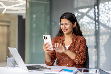 Young woman feeling confused and annoyed while struggling with her mobile phone, looking at the screen with a disappointed expression, sitting at her modern office desk with a laptop