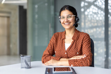 Young woman wearing a headset and glasses sitting at a desk with arms crossed, smiling at the camera, providing professional customer service and support in a modern office environment