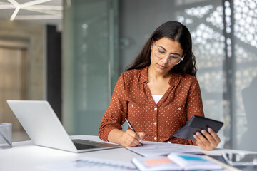 Young businesswoman calculating finances on a calculator, writing notes on documents, and working on a laptop at an office desk, representing accounting, professionalism, and concentrated work