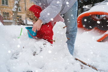 Child in bright red coat and parent enjoy winter by building a small snowman in a snowy playground....