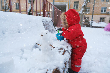 A child in a red snowsuit joyfully builds a small snowman during a snowy day outside. The child...